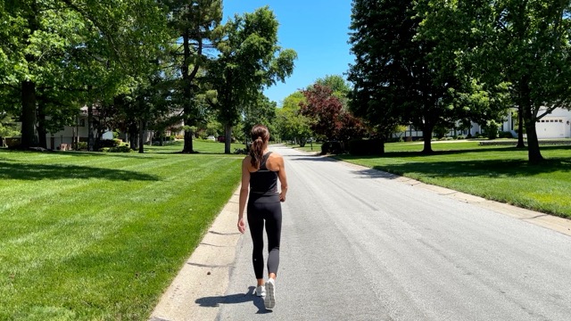 A woman with brown hair tied back, wearing a black tank top and black leggings, walking on the side of a quiet residential street with green trees and grass on a sunny day.
